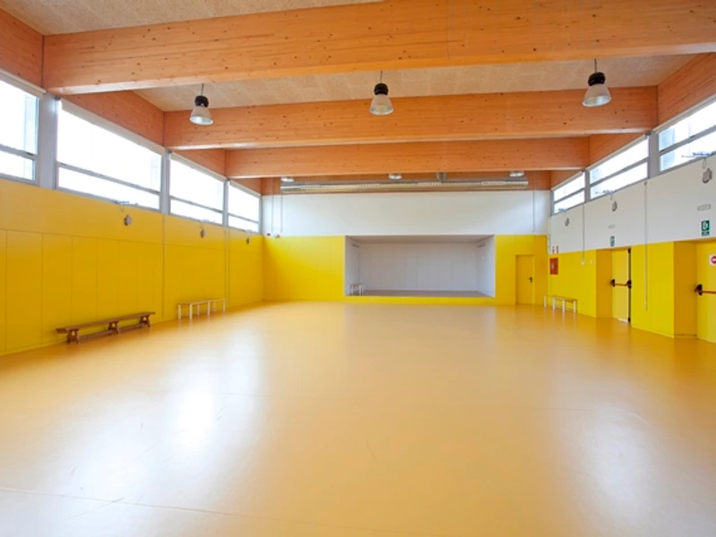 Gimnàs de l’escola, amb terra groc, finestres altes i sostre de fusta.