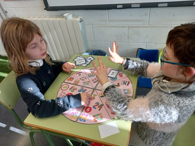 Dues alumnes participen en un joc matemàtic amb una roda i materials manipulatius.